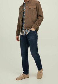 Giacca in suede marrone con due tasche sul petto, indossata sopra una camicia a quadri blu, abbinata a jeans blu scuro e scarpe in suede beige.