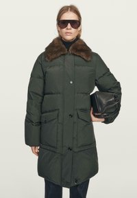 Woman wearing dark green puffer coat with brown fur collar, large sunglasses, holding a black leather handbag against a plain background.