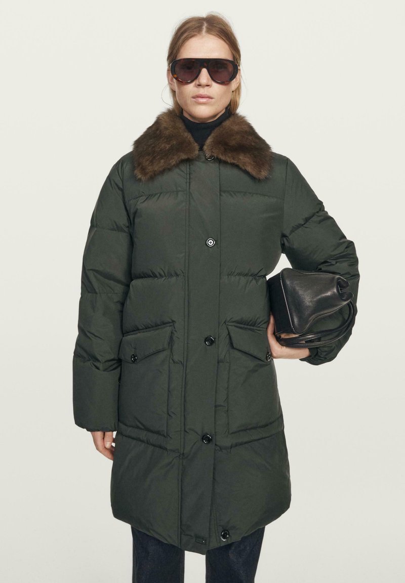 Woman wearing dark green puffer coat with brown fur collar, large sunglasses, holding a black leather handbag against a plain background.