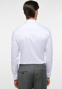Weißes langärmliges Hemd mit strukturiertem Kragen, geknöpften Manschetten und glatter Textur, kombiniert mit grauen Hosen, die über eine Rückentasche verfügen.