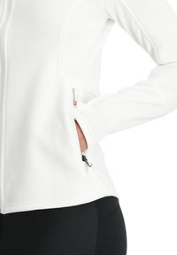 Veste blanche zippée en tissu texturé, dotée de poches latérales, d'un design ajusté et de sections lisses contrastantes le long des manches.