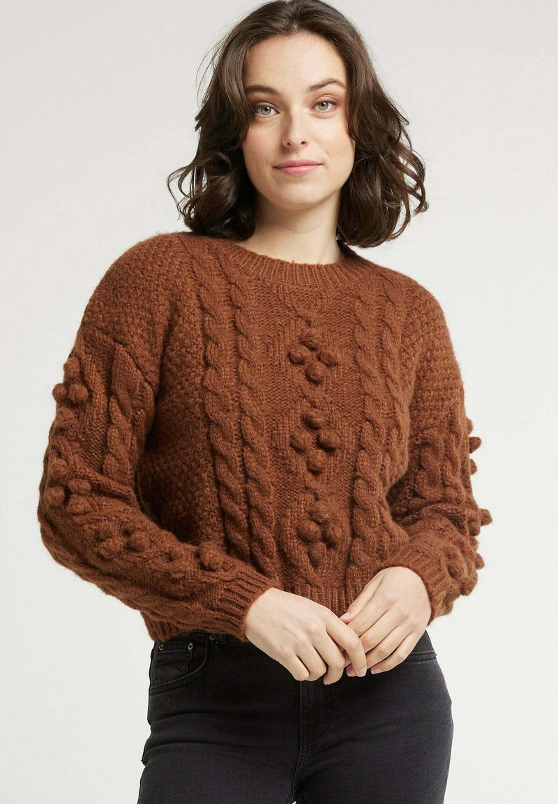 Pull en maille marron avec un design en câble texturé, mettant en avant des accents de boules sur les manches ainsi qu'un col et un ourlet côtelés.