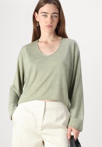 Långärmad, kort topp i mjukt grönt tyg med en vid halsringning och avslappnad passform. Tillsammans med krämfärgade byxor, vilket ger en avslappnad look.