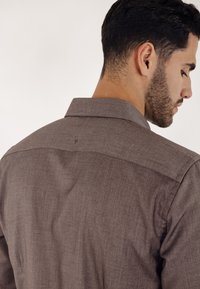 Brun knappeskjorte for menn, teksturert stoff, synlig krage og detaljert søm på ryggen, med lett avrundet kant og synlige sømmer.