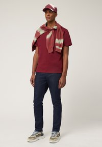 T-shirt color bordeaux, sciarpa tinta unita rossa e verde, jeans in denim scuro, sneakers beige con accenti grigi e cappellino abbinato con dettagli bianchi.