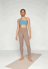 Γυναίκα που στέκεται ξυπόλητη σε ένα στρώμα γιόγκα, φορώντας ένα μπλε αθλητικό σουτιέν με έναν ώμο και ψηλόμεσα μπεζ κολάν σε ουδέτερο φόντο.