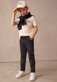 Vit keps med flagga, beige polo tröja, svart tröja över axlarna, mörka byxor, vita sneakers. Enfärgad bakgrund.