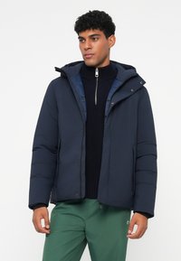 Marineblå pufferjakke med hætte, lynlås og trykknapper. Har et quiltet mønster og elastiske manchetter. Bæres over en mørk sweater.