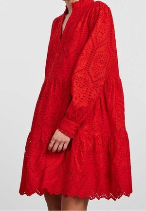 Femme portant une robe rouge à manches longues avec motif en dentelle ajourée et ourlet festonné, montrée du cou au milieu de la cuisse, main reposant sur le côté.