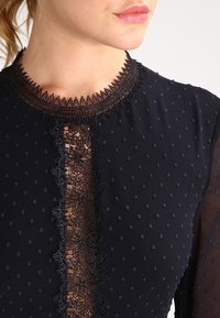 Kleid in Marineblau mit transparenter, gepunkteter Textur und aufwendigen schwarzen Spitzenakzenten am Ausschnitt und in der Mitte, die einen geschichteten Effekt erzeugen.