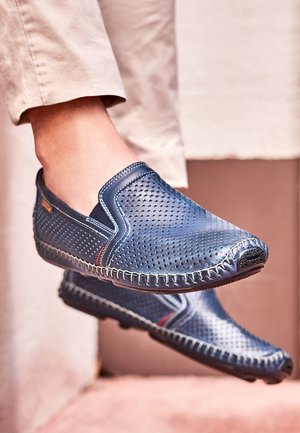 Voeten met blauwe geperforeerde leren instapsloffen met witte stiksels, beige broek, staand tegen een neutrale muur.