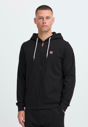 Ung mann med kort hår iført en svart zip-up hettegenser med hvite snører og matchende svarte bukser, stående mot en enkel lys bakgrunn.