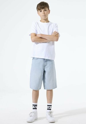 Ragazzo in piedi con le braccia incrociate, indossa una maglietta bianca, pantaloncini azzurri fino al ginocchio, calzini bianchi con motivo e sneakers bianche su sfondo bianco.