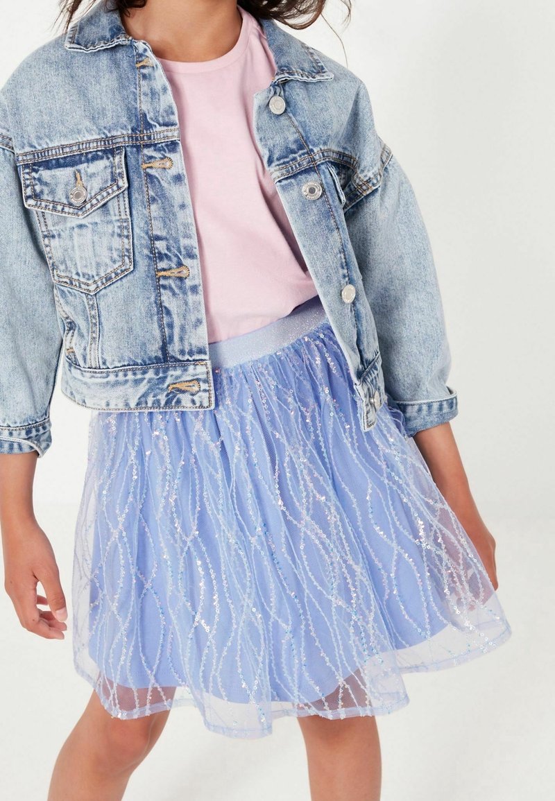 Enfant portant une veste en jean bleu clair, un t-shirt rose pâle et une jupe en tulle bleue scintillante, debout sur un fond blanc uni.