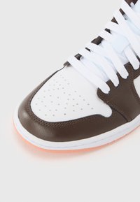 Gros plan sur l'avant d'une basket blanche et marron avec des perforations, des lacets blancs et un bord de semelle orange clair sur un fond uni.