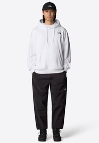 Sudadera blanca con capucha y bolsillo frontal, adornada con un logo negro, combinada con pantalones cargo negros y botas de cordones negras. Una gorra negra completa el conjunto.