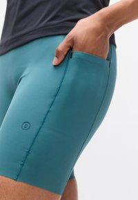 Τιρκουάζ αθλητικό σορτς από ελαστικό ύφασμα, με πλαϊνή τσέπη και επίπεδες ραφές. Συνδυάζονται με σκούρο πουκάμισο.