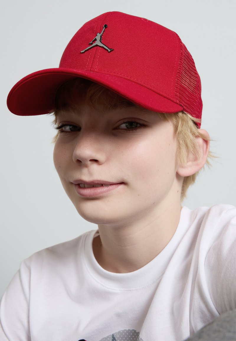 Sarkans cepure ar plakanu malu, kurai ir melns Jordana logotips. Izgatavota no strukturētas auduma ar sietiņa sānu paneļiem, lai nodrošinātu gaisa caurlaidību. Redzama balta T-kreklu.