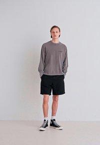 Graues Langarmshirt, schwarze Shorts, weiße Socken und schwarze Sneaker. Der Stoff scheint weich zu sein; das Shirt hat ein kleines Logo auf der Vorderseite. Neutraler Hintergrund.
