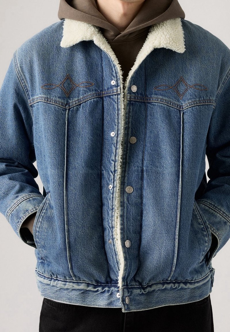 Personne portant une veste en jean bleue avec une doublure en polaire blanche et un sweat à capuche marron en dessous, les mains dans les poches de la veste.