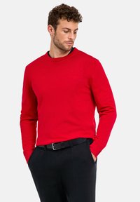 Pull en tricot rouge à col rond avec des manches longues, des poignets côtelés et un tissu texturé. Associé avec un pantalon noir et une ceinture noire.