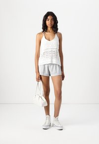 Hvit heklet tanktop med løs passform og intrikat mønster, sammen med lysegrå stripete shorts. Modellen holder en liten hvit håndveske.