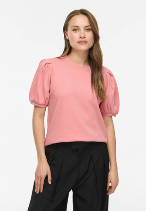 Donna che indossa una camicetta rosa a maniche corte a sbuffo e pantaloni neri plissettati a vita alta, in piedi su uno sfondo bianco.