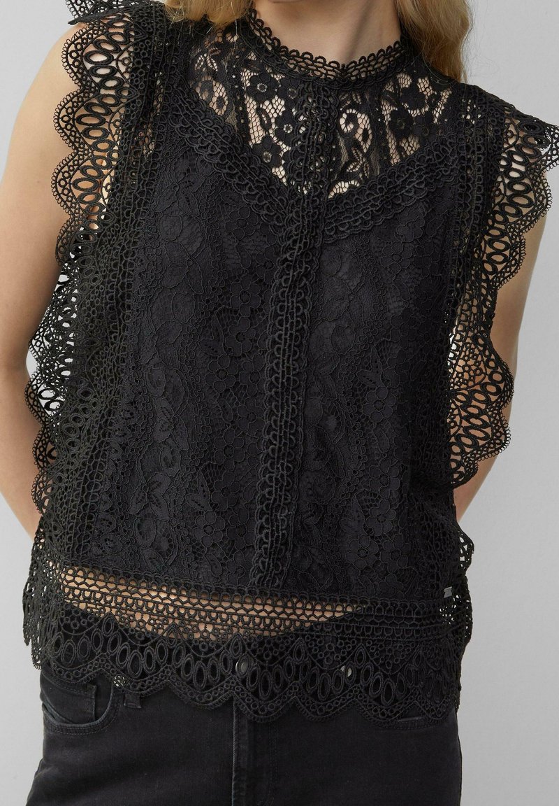 Haut en dentelle noir sans manches avec motifs floraux, bords festonnés et col montant, porté sur un pantalon noir.