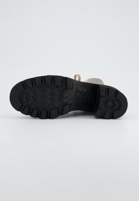 Semelle en caoutchouc noir avec un motif de grip texturé, présentant des formes en zigzag et géométriques ; dessus gris clair partiellement visible.