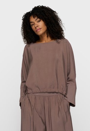 Frau mit lockigen Haaren, die ein locker sitzendes, strukturiertes, mauvefarbenes Langarmoberteil und passende, weit geschnittene Hosen trägt, Hände in den Taschen, neutraler Gesichtsausdruck.
