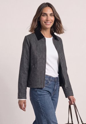 Femme portant une veste à chevrons grise avec un col noir, une chemise blanche, un jean bleu, souriante et tenant les anses d’un sac à main marron dans sa main droite.