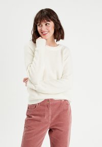 Pull blanc, doux et moelleux avec un col rond, accompagné d'un pantalon en velours côtelé rose comportant des poches avant et un bouton doré.