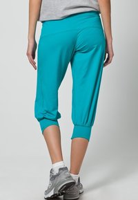 Pantalons turquoise coupés avec une texture lisse, une taille élastique et des poignets ajustés. Portés avec des baskets grises.