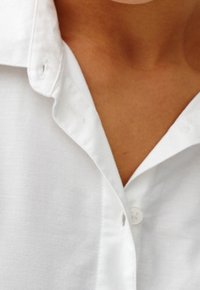 Chemise blanche à boutons avec col, présentant une texture lisse et un bouton unique près du décolleté. Lignes épurées et design classique.