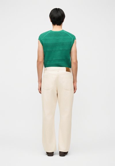 Άτομο με κοντά μαύρα μαλλιά που φοράει πράσινο πλεκτό τοπάκι χωρίς μανίκια και κρεμ παντελόνι υψηλής μέσης με φαρδιά πόδια, γυρισμένο πλάτη σε λευκό φόντο.