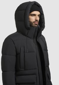 Black padded puffer jacket with a large hood, front pockets, and a zip closure. Features smooth texture and adjustable drawstring details.
