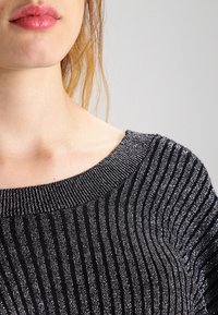 Svart och silverrandig stickad topp med vid halsringning. Materialet verkar mjukt med en glänsande textur från metalltrådsdetaljer.