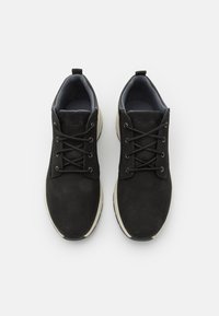 Svarta mockaskor med grått tygfoder, platta snören och vita gummisulor. Har en framträdande logotyp på tungan.