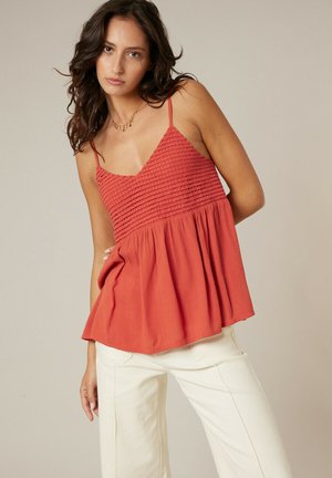 Femme aux cheveux ondulés portant un haut sans manches texturé couleur rouille et un pantalon blanc cassé, debout devant un fond beige uni.