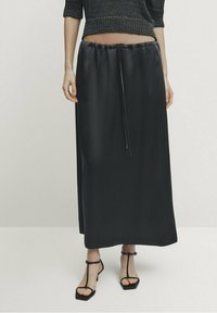 Jupe longue en satin noir avec taille à cordon. Texture lisse avec une silhouette droite, portée avec un haut en maille noir ajusté et des sandales.