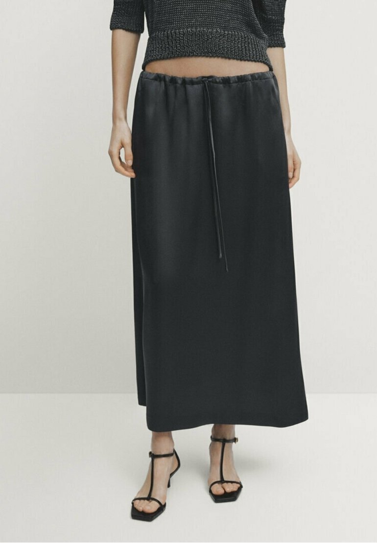 Jupe longue en satin noir avec taille à cordon. Texture lisse avec une silhouette droite, portée avec un haut en maille noir ajusté et des sandales.