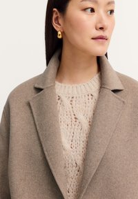 Tanfarbener Wollmantel mit strukturierten Revers über einem cremefarbenen Zopfmustersweater. Das Model trägt goldene, oval geformte Ohrringe. Minimalistisches und klares Design.