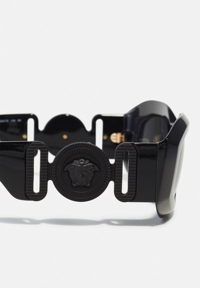 Ochelari de soare din rășină neagră cu un cadru larg, având un emblemă circulară proeminentă a Medusei și detalii distinctive în partea laterală tăiate.