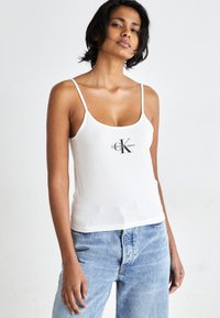 Hvit tanktopp med tynne stropper, med et sentrert svart Calvin Klein-logo, parret med lyseblå jeans. Glatt stoff, uformell design.
