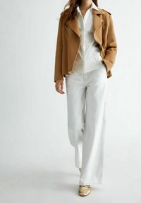 Brązowa dopasowana wełniana kurtka z detalami na zamek, noszona na białej koszuli, zestawiona z białymi, szerokimi spodniami dżinsowymi i jasnobeżowymi butami.