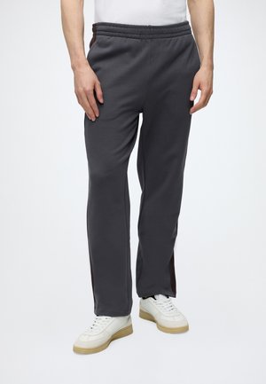 Uomo con pantaloni della tuta grigio scuro e scarpe da ginnastica bianche su sfondo chiaro uniforme, mani rilassate ai lati.