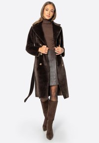 Abrigo de piel sintética marrón de doble botonadura con grandes solapas, botones dorados y un cuello de tortuga marrón a juego. Falda lápiz de cuero gris. Botas marrones hasta la rodilla.