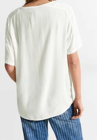 Vit blus med korta ärmar och avslappnad passform. Har ett mjukt, lätt material och rundad fåll. Bärs med blå randiga jeans.