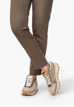 Sneakers with a beige suede upper, white laces, gold accents, and a chunky beige sole. Paired with fitted brown pants, showcasing a casual style.