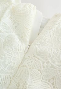 Detailný záber bielej tkaniny s kvetinovou čipkou a viditeľným bielym zipsom na odeve.